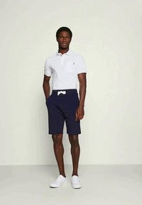 Balts polo krekls ar mazu logo, tumši zili šorti ar auklu, un baltas sporta kurpes. Audums šķiet gluds un neformāls.