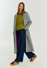 Šedý vzorovaný kabát přes zelený pletený svetr, doplněný širokými tmavými džíny a hnědými teniskami s bílými pruhy a gumovými podrážkami.