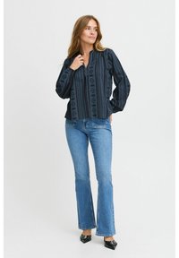 Blusa a maniche lunghe di colore blu scuro con motivo geometrico e scollo a V, abbinata a jeans a campana di colore blu chiaro e tacchi neri. Il design presenta stampe geometriche.