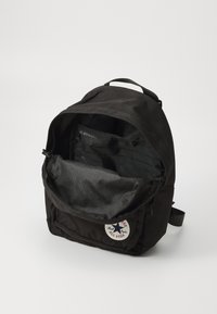 Zaino in tessuto nero con scomparto principale con zip, tasca interna e una etichetta bianca con il logo Converse. Superficie texturizzata.