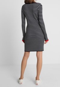 Schwarz-weiß gestreiftes, geripptes Kleid mit langen Ärmeln und roten Manschetten. Das Kleid ist figurbetont und fällt über das Knie.