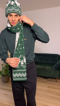 Bonnet en tricot vert et écharpe avec motifs de rennes blancs et d'arbres. Texture douce, écharpe rectangulaire drapée autour du cou. Pantalon noir.