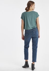 Top corto de manga corta verde con un corte relajado, combinado con jeans oscuros de corte ajustado. El modelo lleva botas negras tipo tobillo. Fondo liso.
