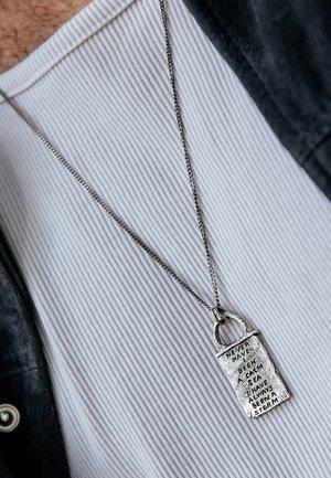 Collier en pendentif argenté de forme rectangulaire avec du texte gravé. La chaîne est fine et métallique, contrastant avec une chemise blanche à côtes.