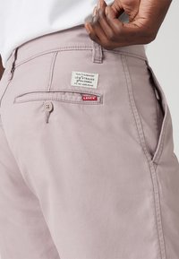 Pantaloni Levi's de un roz deschis, cu un buzunar din spate vizibil, prezentând un eticheta. Materialul are o textură netedă și o croială clasică.