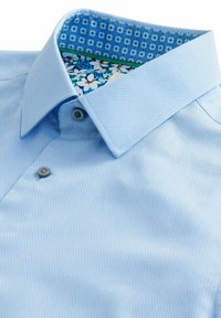 Polo shirt bleu clair en tissu texturé, présentant un imprimé floral contrastant à l'intérieur du col et deux boutons sur le rabat.