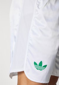 Baltieji sportiniai šortai, pagaminti iš lygaus audinio, su žaliu „Adidas“ logotipu kairėje kojoje, su siaurėjančiu kojos dizainu ir šoninėmis siūlėmis.