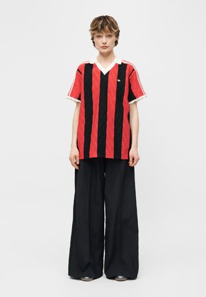 Person trägt ein rotes und schwarzes Hemd mit vertikalen Streifen, weißem Kragen und schwarze weit geschnittene Hosen, steht vor weißem Hintergrund.