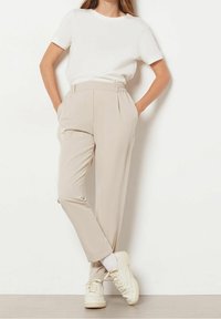 Pantalon beige taille haute avec plis, poches et jambe fuselée. Associé à un T-shirt blanc à col rond et des baskets claires.