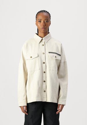 Cremefarbendes, übergroßes Denimhemd mit Kragen, zwei Fronttaschen und Knopfverschluss, das schwarze Akzentnähte an einer Tasche aufweist.
