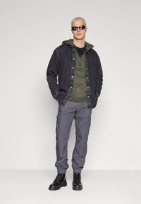 Codzienny strój składający się z ciemnej kurtki dżinsowej na oliwkowej zielonej bluzie z kapturem, w połączeniu z szarymi spodniami typu cargo i czarnymi sznurowanymi butami.