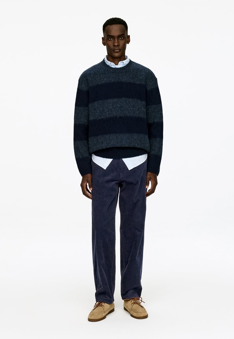 Un pull rayé bleu marine en matériau doux, associé à une chemise à col bleu clair, un pantalon en velours côtelé noir et des chaussures beige à lacets.
