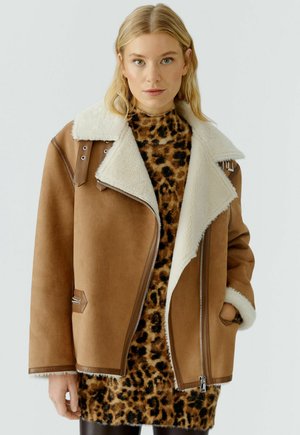 Braune Fake-Suede-Jacke mit einem weißen Schaffellkragen, Reißverschluss und metallischen Details, getragen über einem Kleid mit Leopardenmuster.
