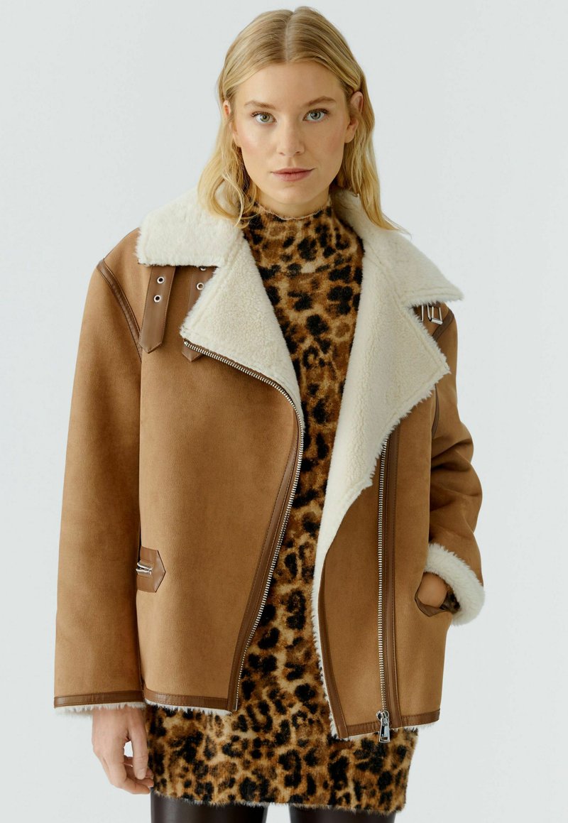 Veste en suédine marron avec un col en fourrure synthétique blanche, fermeture éclair et détails métalliques, portée sur une robe pull à imprimé léopard.