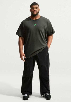 Uomo in piedi che indossa una t-shirt Nike grigio scuro, pantaloni neri a gamba larga e scarpe da ginnastica nere su uno sfondo chiaro uniforme.