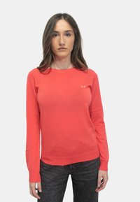 Maglione a manica lunga corallo realizzato in morbido tessuto a maglia, caratterizzato da un collo rotondo e un piccolo logo sul petto, abbinato a jeans scuri.
