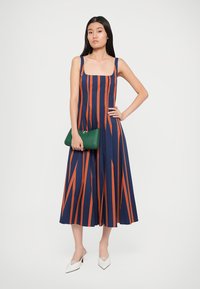 Γυναίκα που φοράει ένα άσπρο αμάνικο ναυτικό φόρεμα με πορτοκαλί κατακόρυφες ρίγες, κρατώντας μια πράσινη τσάντα και φορώντας λευκά μυτερά τακούνια.