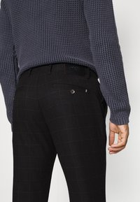 Szary sweter z dzianiny o fakturowanym wzorze, zestawiony z czarnymi spodniami w kratę, posiadającymi tylną kieszeń i kontrastujący paskowany detal.