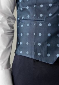 Navyblauer Polka-Dot-Weste aus Stoff mit hellblauen Punkten, mit schlanker Passform und Tascheninfo, kombiniert mit einem weißen Hemdärmel und dunklen Hosen.