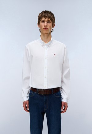 Mladý muž nosící bílou košili s knoflíky a malou vlajkou jako znakem, modré džíny a hnědý pásek, stojící před světlým pozadím.