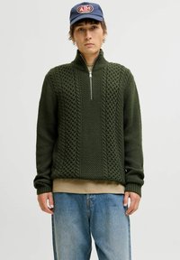 Pull en maille vert foncé avec un col haut, une fermeture éclair quart, un motif tressé complexe et des poignets côtelés, superposé sur une chemise beige et un jean bleu.