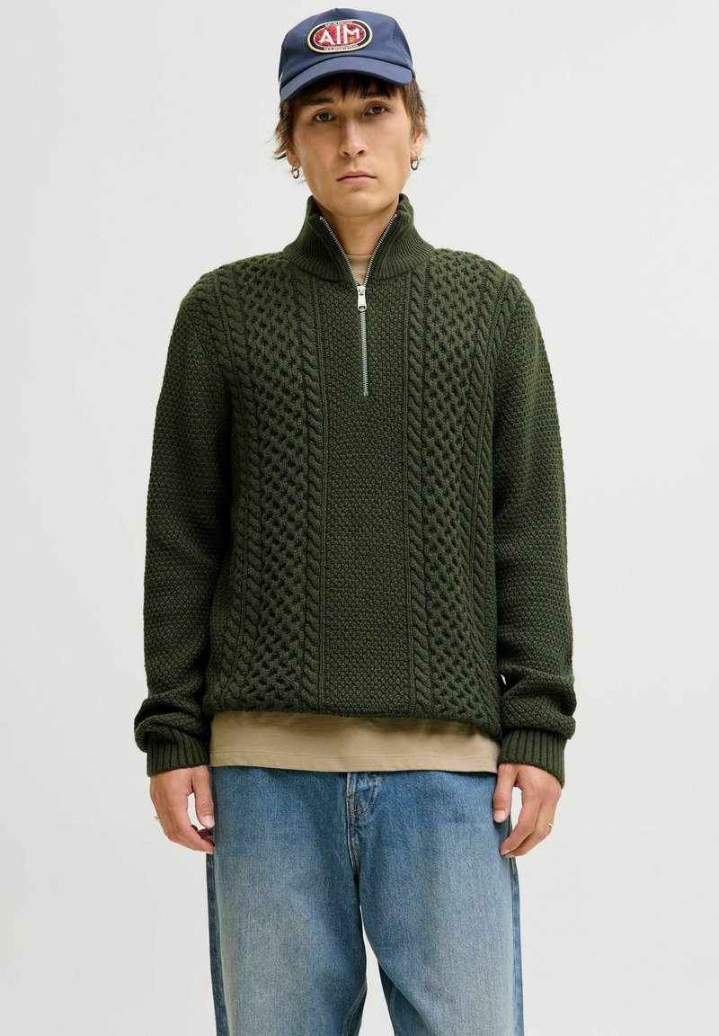 Pull en maille vert foncé avec un col haut, une fermeture éclair quart, un motif tressé complexe et des poignets côtelés, superposé sur une chemise beige et un jean bleu.
