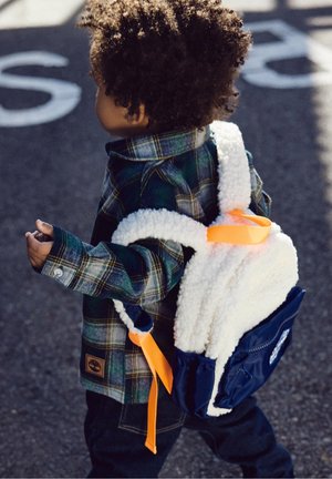 Kleinkind mit lockigem Haar läuft auf Asphalt, trägt eine karierte Jacke und einen weißen und marineblauen Rucksack mit orangefarbenen Riemen.