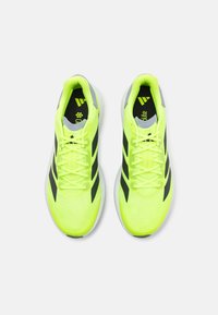 Neon yellow athletic shoes with black accents, mesh upper, padded collar, and flat laces. Features grey heel support and textured sole.