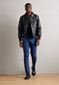 Chaqueta bomber de cuero negro con puños acanalados, llevada sobre una camisa negra. Combinada con pantalones azules ajustados y zapatos negros de cordones.