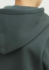 Felpa verde scuro con cappuccio, caratterizzata da una superficie liscia e un design semplice. Il tessuto sembra resistente e leggero.
