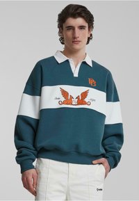 Pulover teal cu dungi albe orizontale, având un design brodat cu două lei cu aripi și logo pe piept. Guler alb.