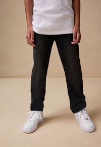Pantalon en denim noir à coupe droite, associé à des baskets blanches. Le haut est un t-shirt blanc ample. Le fond est beige.