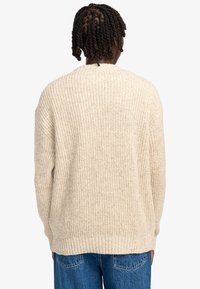 Beige chunky breiseltrui met een losjes vallende pasvorm, dikke ribbels, verlaagde schouders en een brede halslijn. Open rug met zijslits.