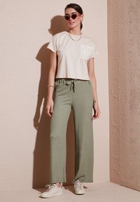 Crèmekleurige crop top met een gestructureerde zak, gecombineerd met losse, ademende, olijfgroene broek. Model draagt witte sneakers en een zonnebril.