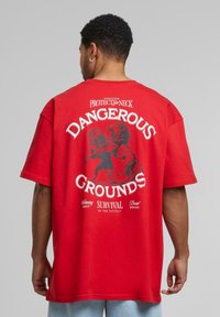 Rød bomulds T-shirt med et stort sort grafisk print på ryggen med teksten: "DANGEROUS GROUNDS" og en løveillustration. Kortærmede, afslappet pasform.