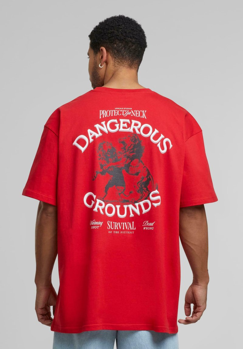 Rød bomull T-skjorte med et stort svart grafisk motiv på baksiden med teksten: "DANGEROUS GROUNDS" og en løveillustrasjon. Korte ermer, avslappet passform.