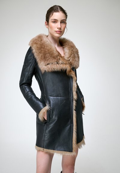 Jeune femme portant un manteau en cuir noir avec un grand col et des poignets en fourrure marron, debout les mains dans les poches devant un fond uni.