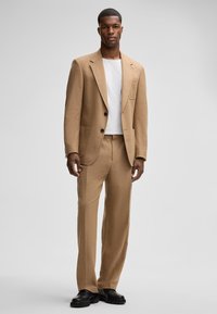 Beżowy garnitur szyty na miarę, wykonany z teksturowanej tkaniny, z jednorzędową marynarką na dwa guziki i dopasowanymi spodniami, noszony z białą koszulą.