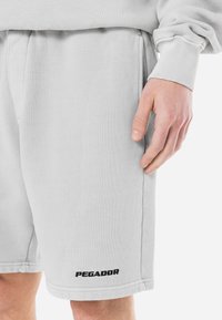 Pantalones cortos de color gris claro hechos de una tela texturizada, con una cinturilla elástica y un logo negro "PEGADOR" cerca del dobladillo.
