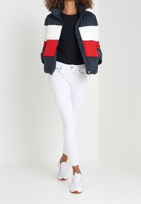 Kvinna som bär en marinblå, vit och röd färgblockeringspufferjacka, svart topp, vita jeans och vita sneakers, står med händerna i fickorna.