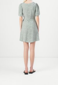 Osoba stojąca tyłem, ubrana w zieloną sukienkę z krótkim rękawem w białe kropki oraz czarne sandały na odkrytej pięcie na białym tle.