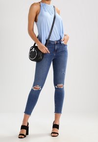 Haut sans manches bleu clair avec détails en dentelle, associé à un jean skinny bleu foncé déchiré et des talons noirs à brides. Petit sac bandoulière noir.