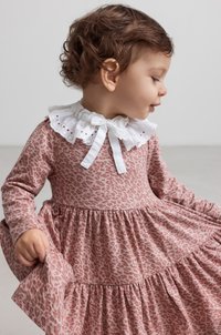 Et ungt barn med krøllet hår har på seg en rosa leopardmønstret kjole med en hvit blonderkrage, mens barnet holder kjolen mens de ser bort.