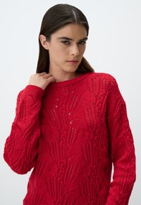 Maglione rosso lavorato a maglia con un motivo testurizzato e lavorazione a giorno, colletto e polsini a costine. Maniche lunghe e materiale morbido.