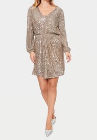 Kort, glittrande klänning med guldpaljetter och V-halsringning samt långa ärmar, med en samlad midja och flödande tyg. Bärs med silvriga klackar.