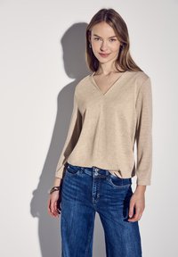 Beige, langärmliges Oberteil mit V-Ausschnitt. Weicher Stoff, lockere Passform. Kombiniert mit dunkelblauen, etwas weiter geschnittenen Jeans. Das Modell steht vor einem hellen Hintergrund.