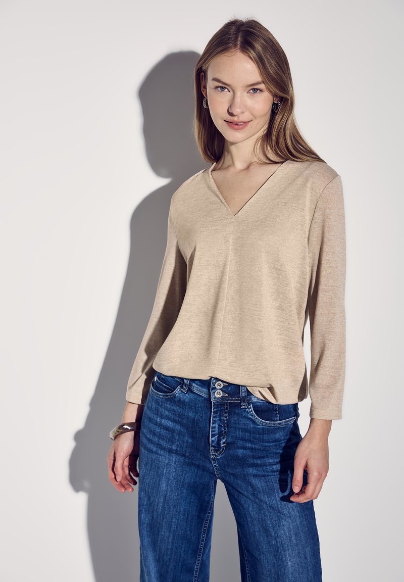 Beige, langärmliges Oberteil mit V-Ausschnitt. Weicher Stoff, lockere Passform. Kombiniert mit dunkelblauen, etwas weiter geschnittenen Jeans. Das Modell steht vor einem hellen Hintergrund.