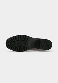 Semelle de botte marron avec une profonde bande de roulement noire présentant des formes de croix et des motifs en blocs pour une bonne traction sur un fond clair uni.