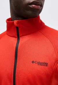 Κοντινό πλάνο ενός ατόμου που φοράει αθλητικό μπουφάν Columbia Titanium με φερμουάρ, σε κόκκινο χρώμα, με υφασμάτινη υφή και ψηλό γιακά.