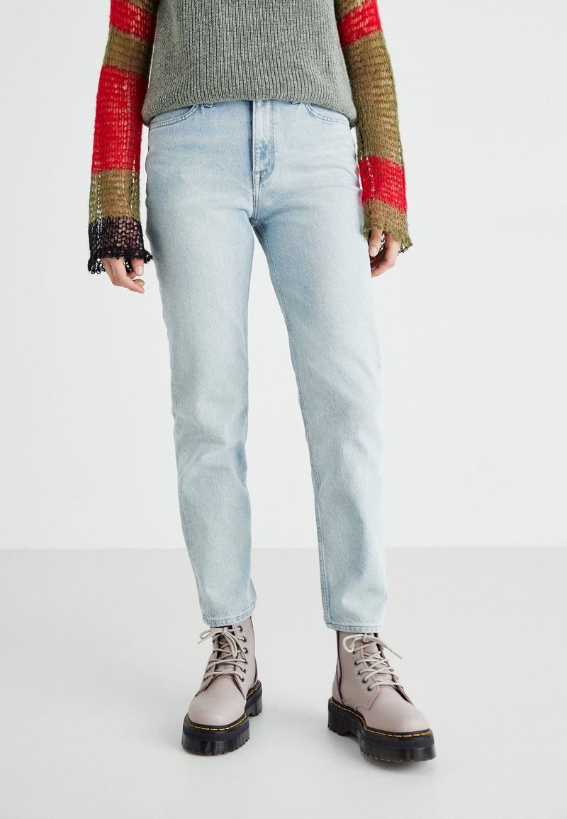 Ljusblå jeans i kombination med en grågrön tröja med randiga ärmar i flera färger, samt grå snörade ankelstövlar med tjocka svarta sulor.